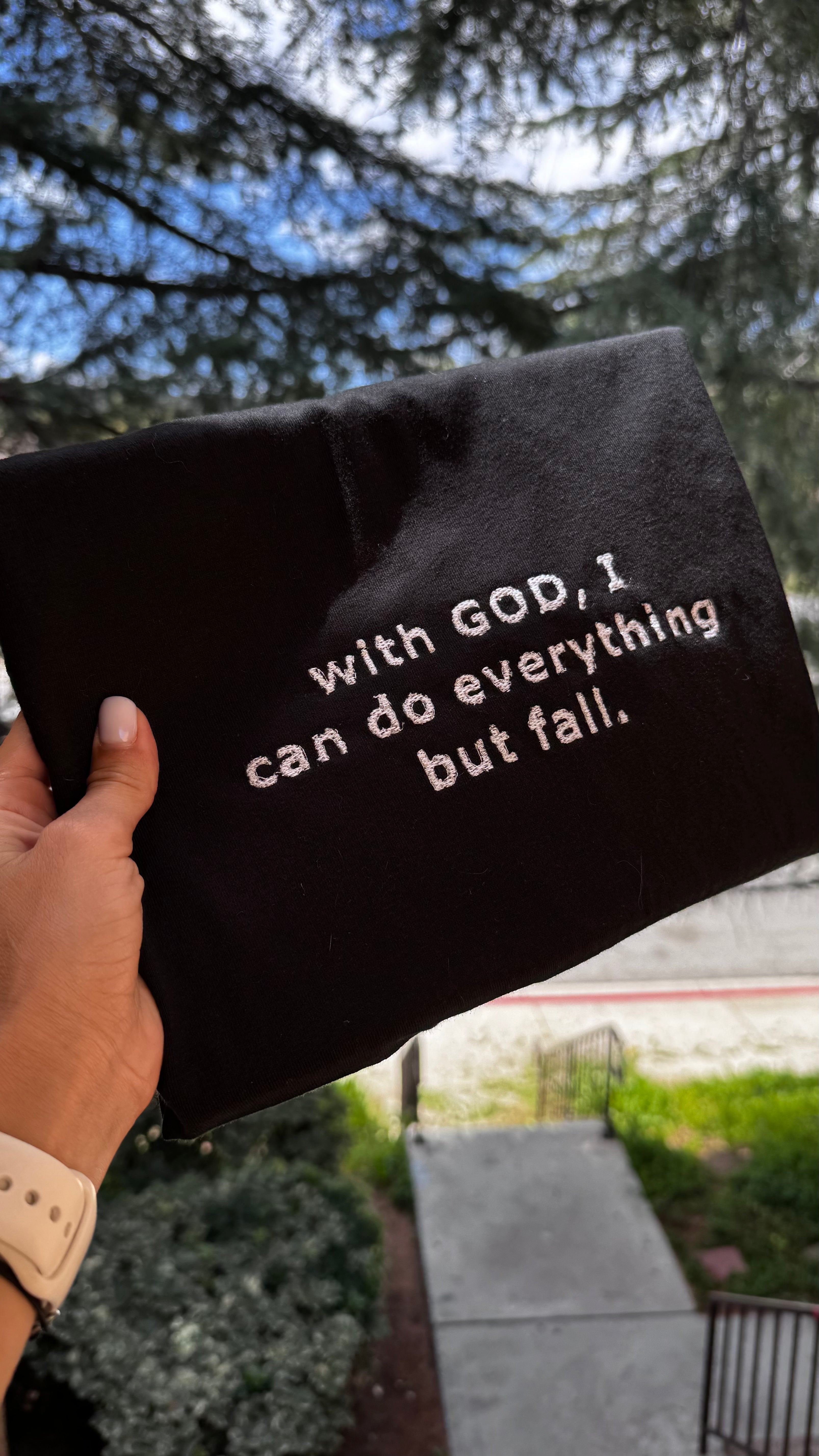 Black inspirational pouch with white Christian quote 'With God, I can do everything but fall' – faith-based accessory by TheALONA, photographed outdoors.
